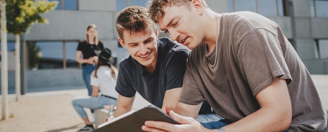 Zwei Studenten sitzen vor der Hochschule und schauen sich Unterlagen 