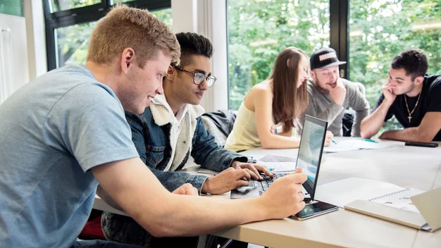 Vier Studierende sitzen an einem Tisch im Freien um einen Laptop herum.