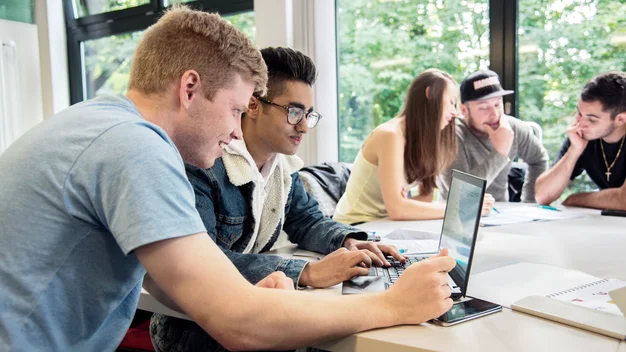 Vier Studierende sitzen an einem Tisch im Freien um einen Laptop herum.