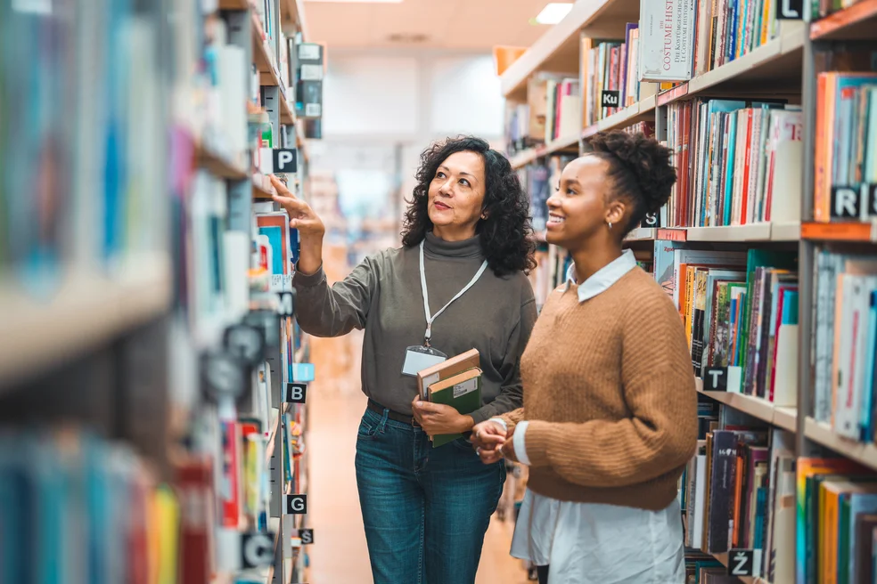 Mid Adult Hispanic Librarian Escorting A Student In A Library
