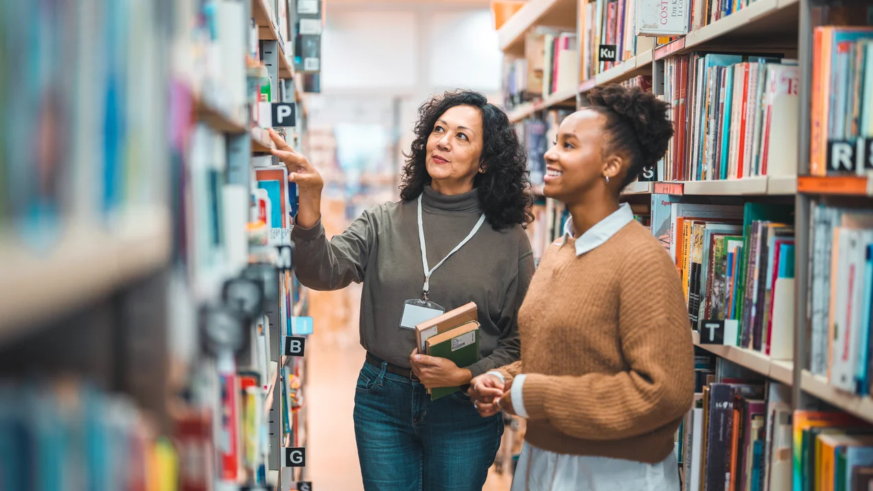 Mid Adult Hispanic Librarian Escorting A Student In A Library