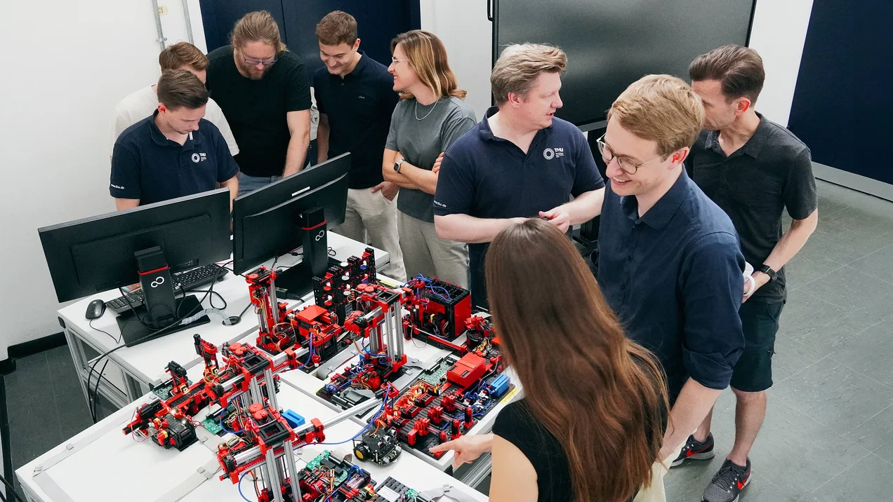 Menschengruppe steht im Kreis um einen Tisch. Auf dem Tisch befindet sich ein Forschungsaufbau zur Industrie 4.0 in Fischertechnik.