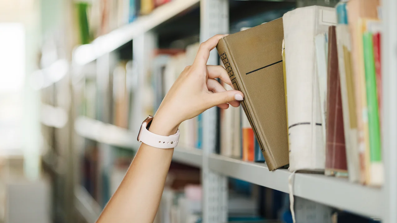 Human hand taking book from shelf in college library. Female took one book. Close-up