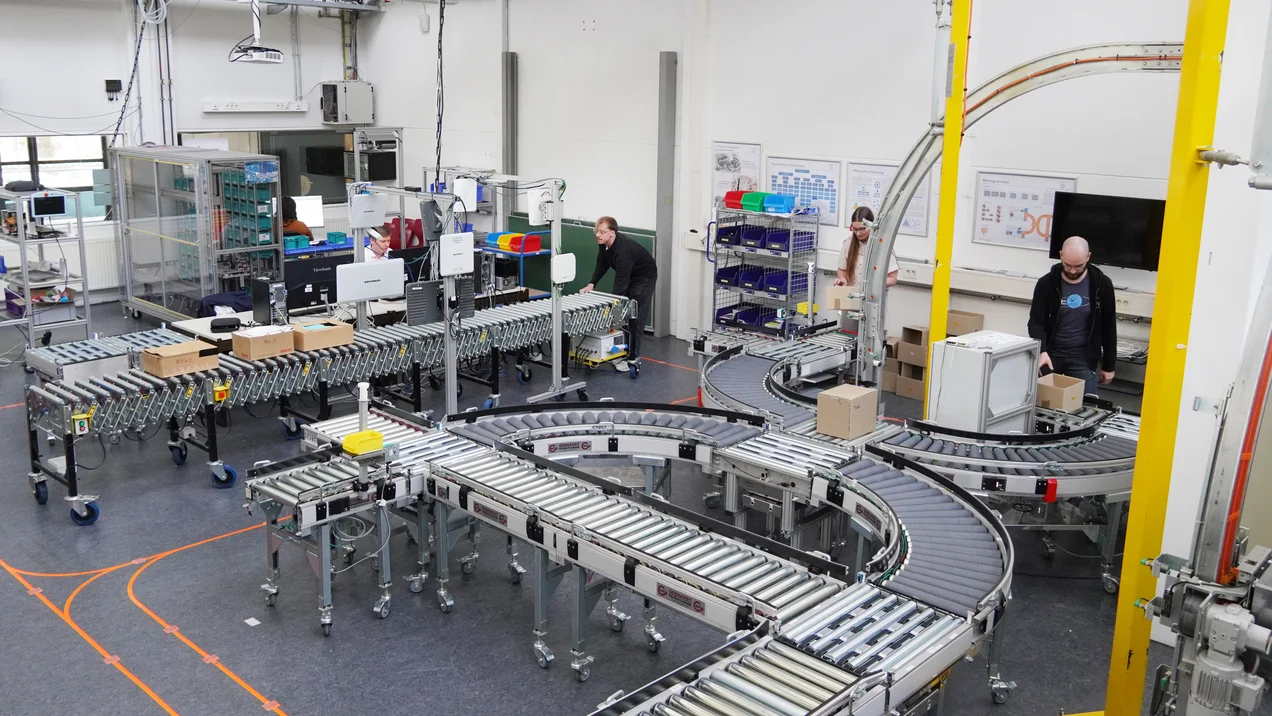 Raumaufnahme der Lernfabrik. Studierende und Mitarbeitende bedienen das flexible Fördersystem FlexFörderer