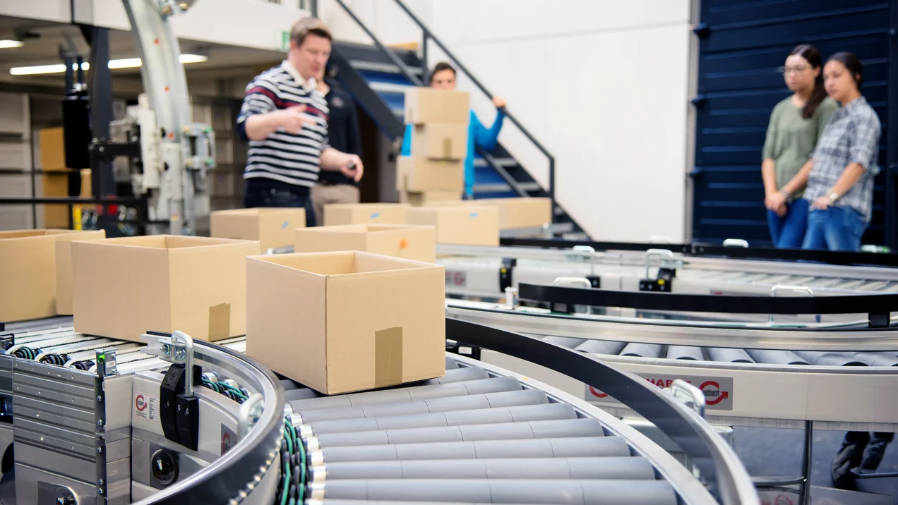 Kartonboxen werden über ein Förderlaufband transportiert. Im Hintergrund erklärt ein Mitarbeiter zwei Studentinnen die Transportvorgänge.