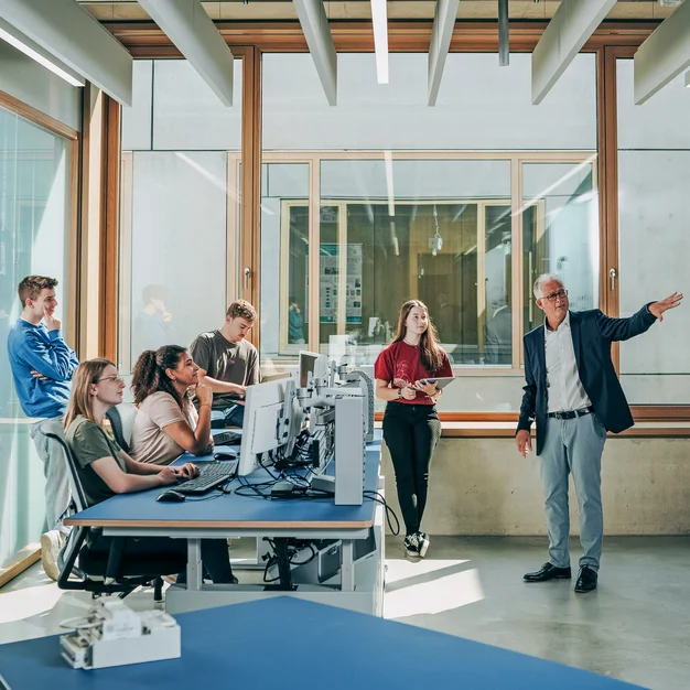 Interaktive Lehrsituation im Smartgrid-Labor: Professor im Dialog mit Studierenden an Computerarbeitsplätzen (Querformat).