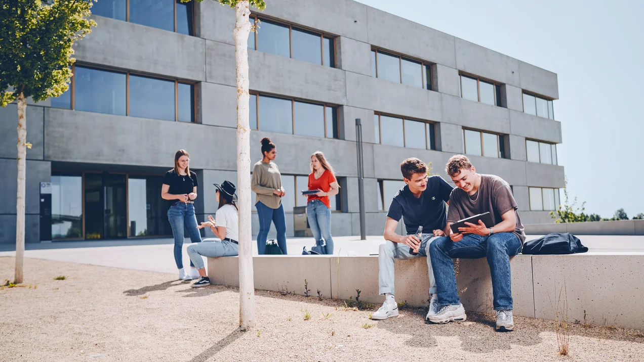 Studierende sitzen entspannt auf einer Betonmauer vor dem modernen Campusgebäude.