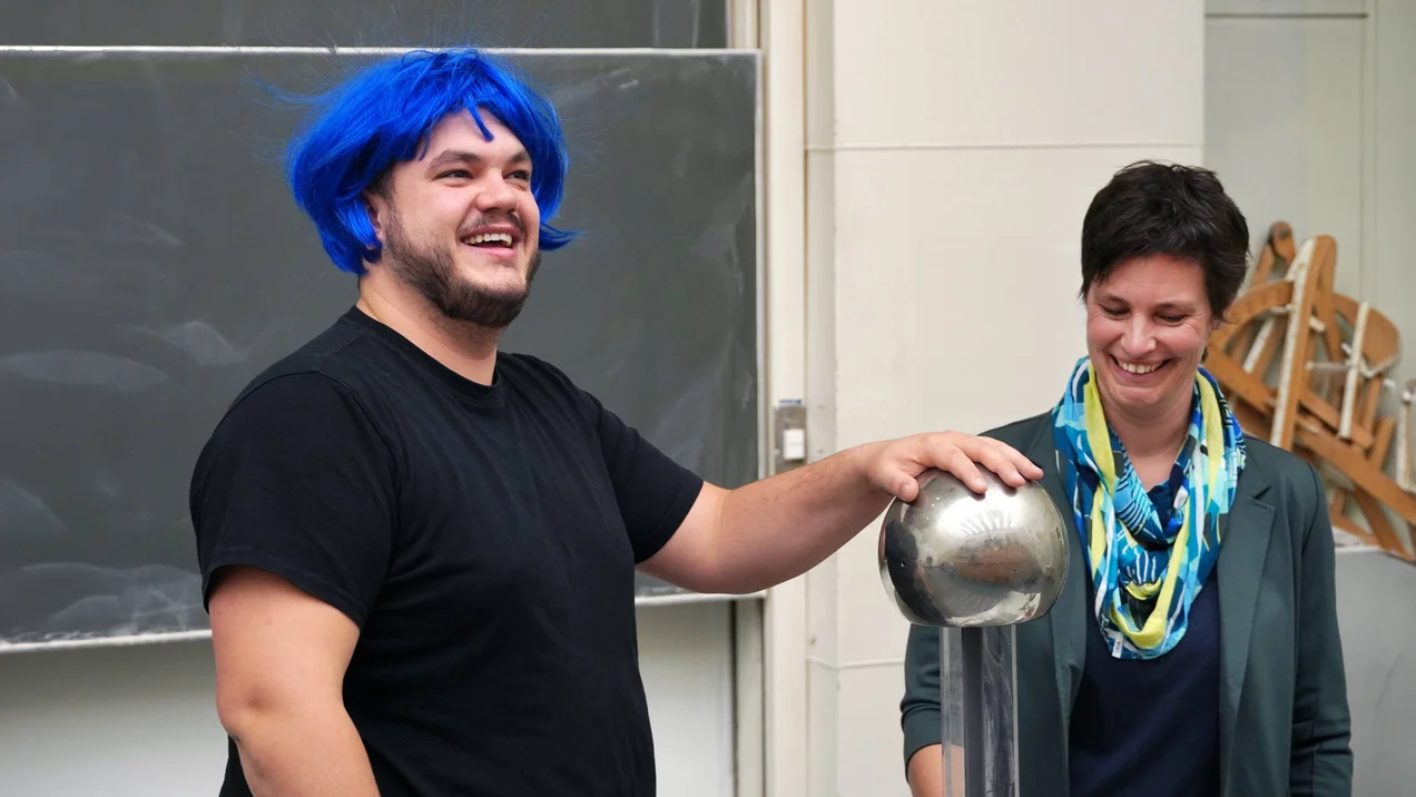 Student und Professorin an einer Boadingkugel von Hubbs. Der Student trägt eine blaue Perücke, die Haare stehen senkrecht nach oben ab.
