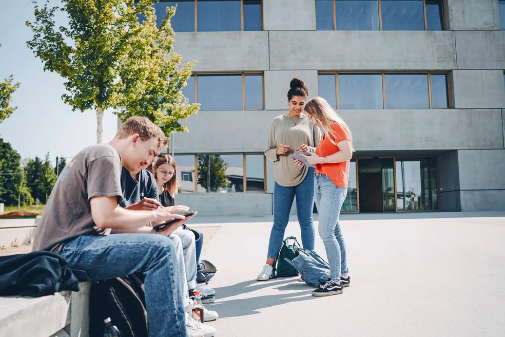 Entspanntes Arbeiten im Freien: Personen mit Laptops auf dem Campusgelände. Gut nutzbar für Flexibilität und moderne Arbeits-/Lernumgebungen.