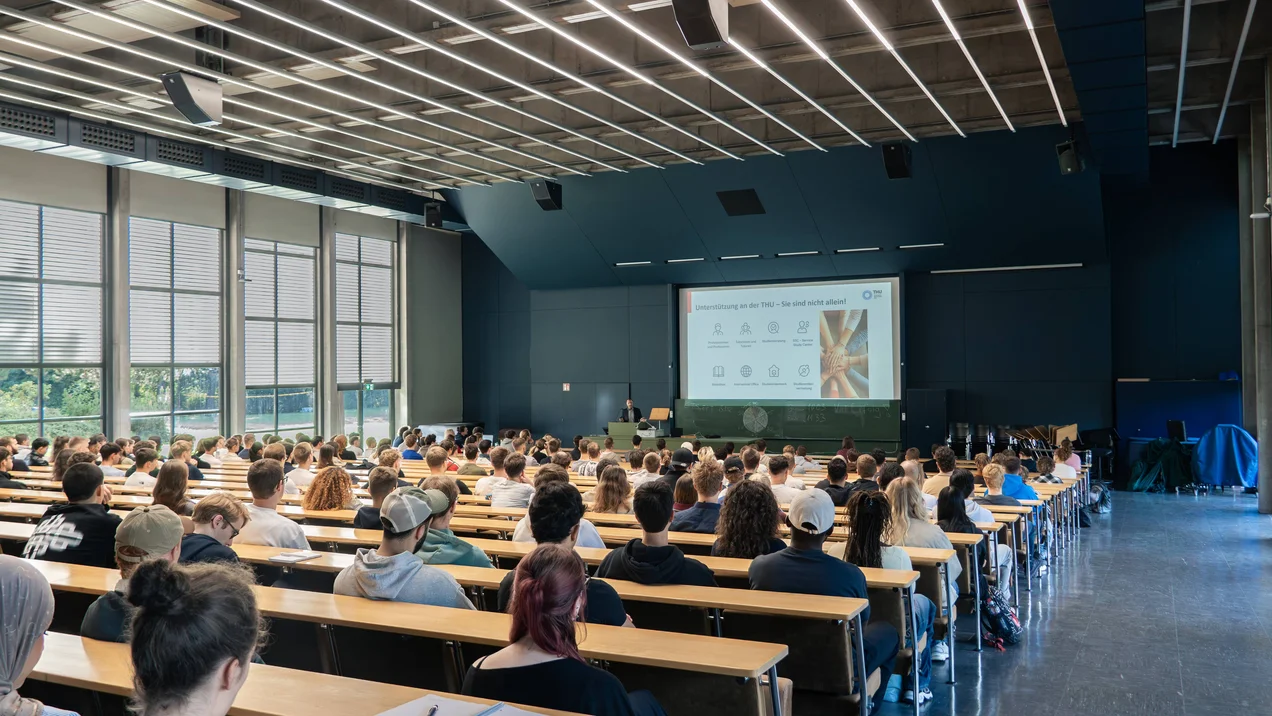 Studenten sitzen in der Aula und werden begrüßt.