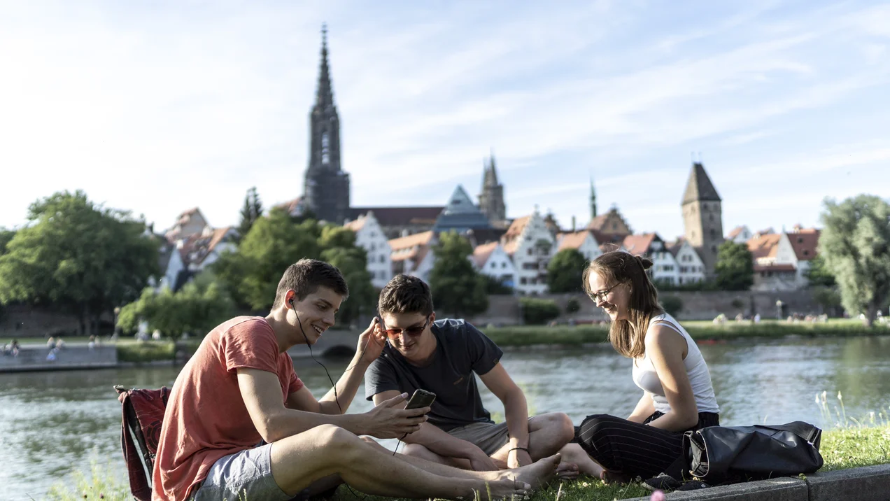 Studierende sitzen entspannt am Donauufer, im Hintergrund die Ulmer Altstadt und das Münster.