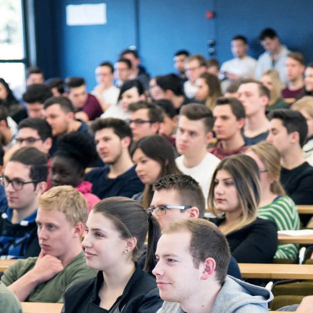 Dicht sitzende Studierende in einem Hörsaal, der Fokus liegt auf den Zuhörern.