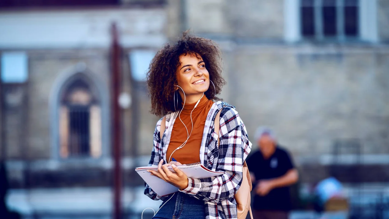 Eine junge Frau mit Notizbuch steht lächelnd vor einem Gebäude auf dem Campus der Technischen Hochschule Ulm und bereitet sich auf ihr Auslandsstudium vor.