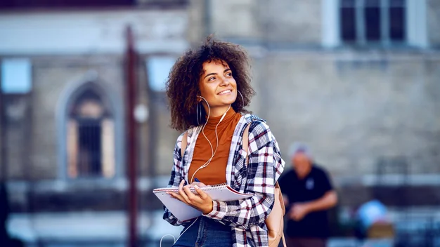A young woman with a notebook stands smiling in front of a building on the campus of Ulm University of Applied Sciences, preparing for her studies abroad.