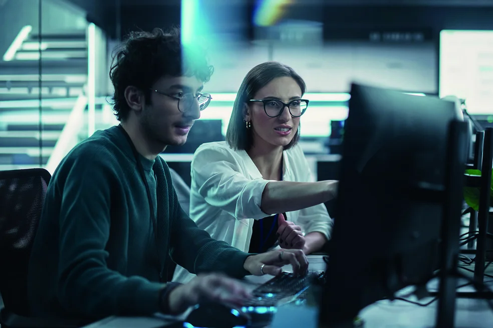 Zwei junge Ingenieure die am Computer arbeiten und sich am Arbeitsplatz unterhalten. Weibliche und männliche Softwareentwickler diskutieren eine Lösung für ihr kollaboratives Projekt zur künstlichen Intelligenz