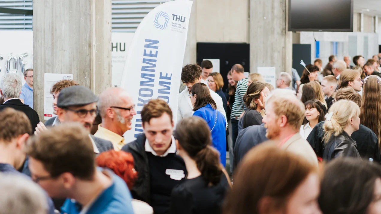 Eine große, feierlich gekleidete Menschenmenge bei einer Veranstaltung, ein Banner im Hintergrund.