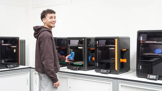 Ein junger Mann lächelt in die Kamera und holt ein kleines Auto aus einem 3D-Drucker