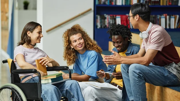 Students are sitting on a bench indoors in front of a bookshelf, chatting. One young woman in the group is in a wheelchair.