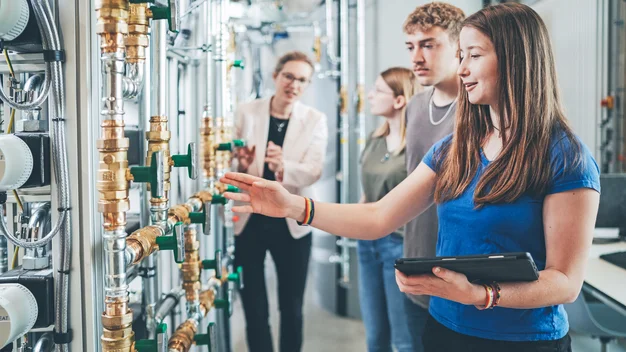 Praxisnahe Lehre im Speicherlabor an der THU: Eine Dozentin erläutert einer Gruppe von Studierenden die Funktionsweise der industriellen Anlage. Im Vordergrund prüft eine Studentin die Rohrleitungen und Ventile, unterstützt durch ein Tablet.