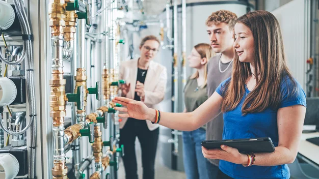 Praxisnahe Lehre im Speicherlabor an der THU: Eine Dozentin erläutert einer Gruppe von Studierenden die Funktionsweise der industriellen Anlage. Im Vordergrund prüft eine Studentin die Rohrleitungen und Ventile, unterstützt durch ein Tablet.