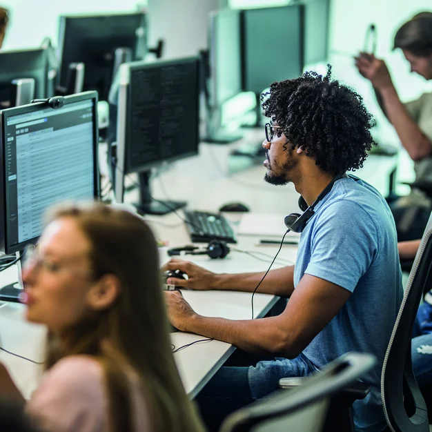 964216144 African American programmer working on desktop PC in the office full of people.