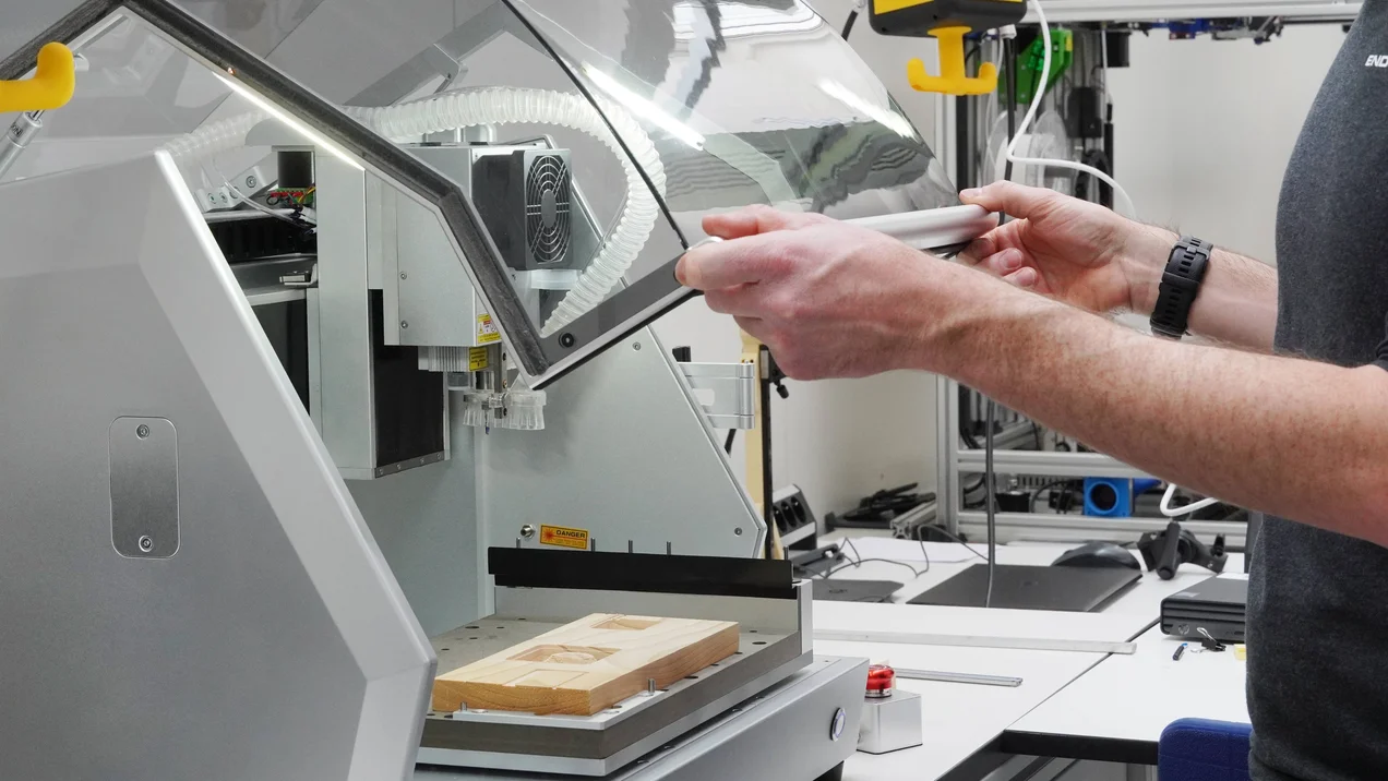 Person öffnet die Schutzhaube einer CNC-Fräsmaschine mit eingespanntem Holzwerkstück in einer Laborumgebung.
