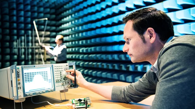 Measurement in the absorber chamber, a room equipped with RF absorbers. A person in the foreground is turning the knob of a measuring device. In the background, a person is working on an antenna.