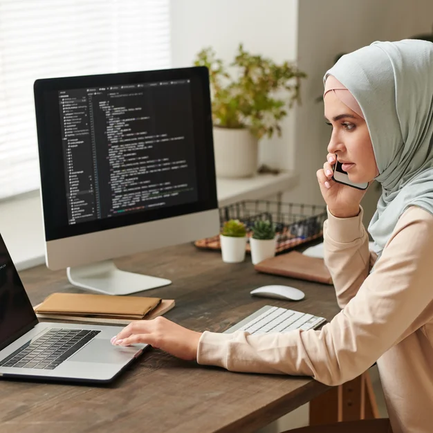 Young woman wearing light blue hijab sitting at table discussing important issues on phone while working on software Young woman wearing light blue hijab sitting at table discussing important issues on phone while working on software