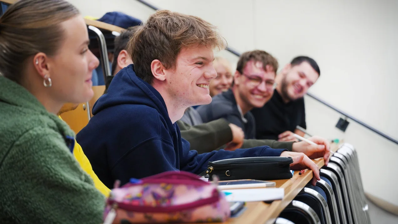 Studierende sitzen lachend in einer Stuhlreihe im Hörsaal bei der Physikvorlesung