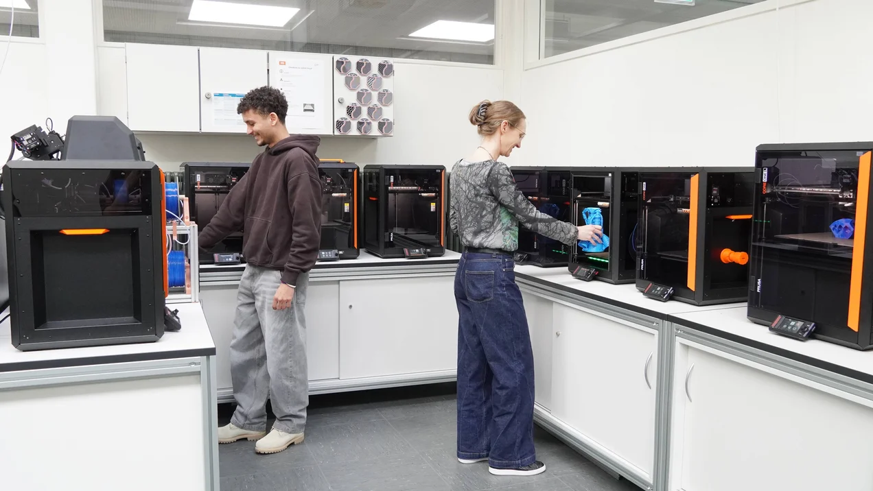 Ein junger Mann und eine Frau bedienen zwei Drucker in einer 3D Drucker Farm